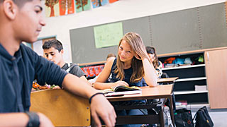 Učenica stara oko 15 godina s otvorenom knjigom na školskoj klupi, kraj nje sjede njezine kolege iz razreda
